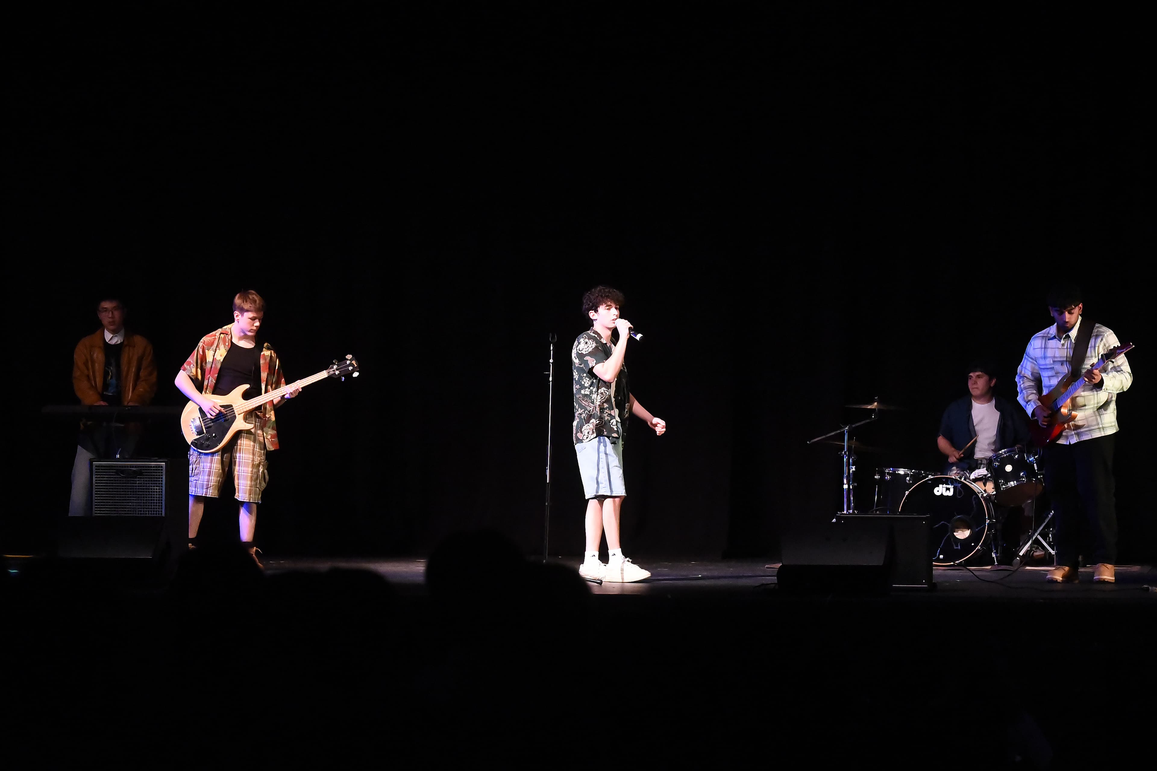 Five student musicians performing on a darkened stage: a vocalist at the microphone, a bassist, a guitarist, a keyboardist, and a drummer.