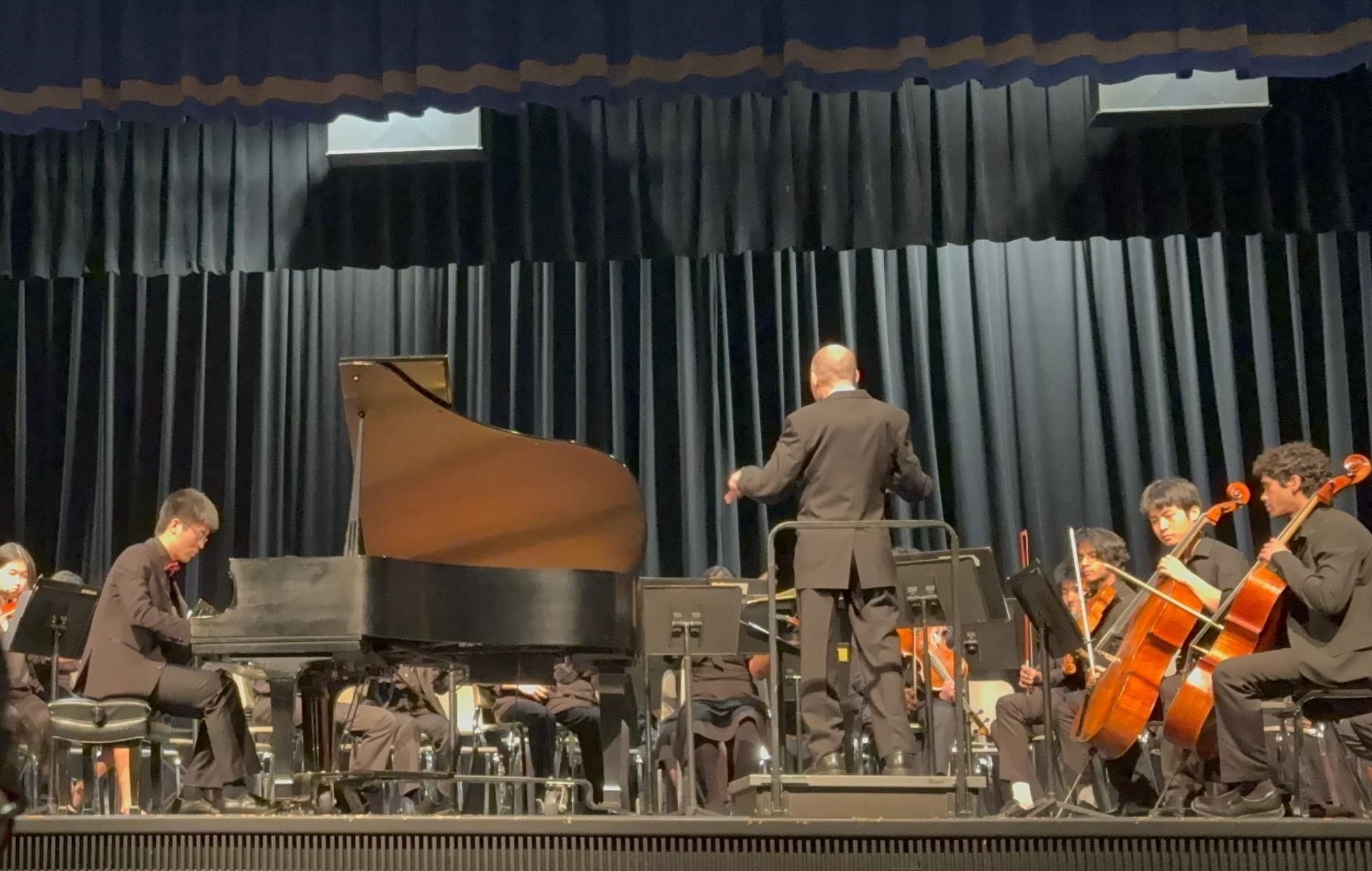 A pianist at a grand piano performing with a school orchestra while a conductor leads from a podium.
