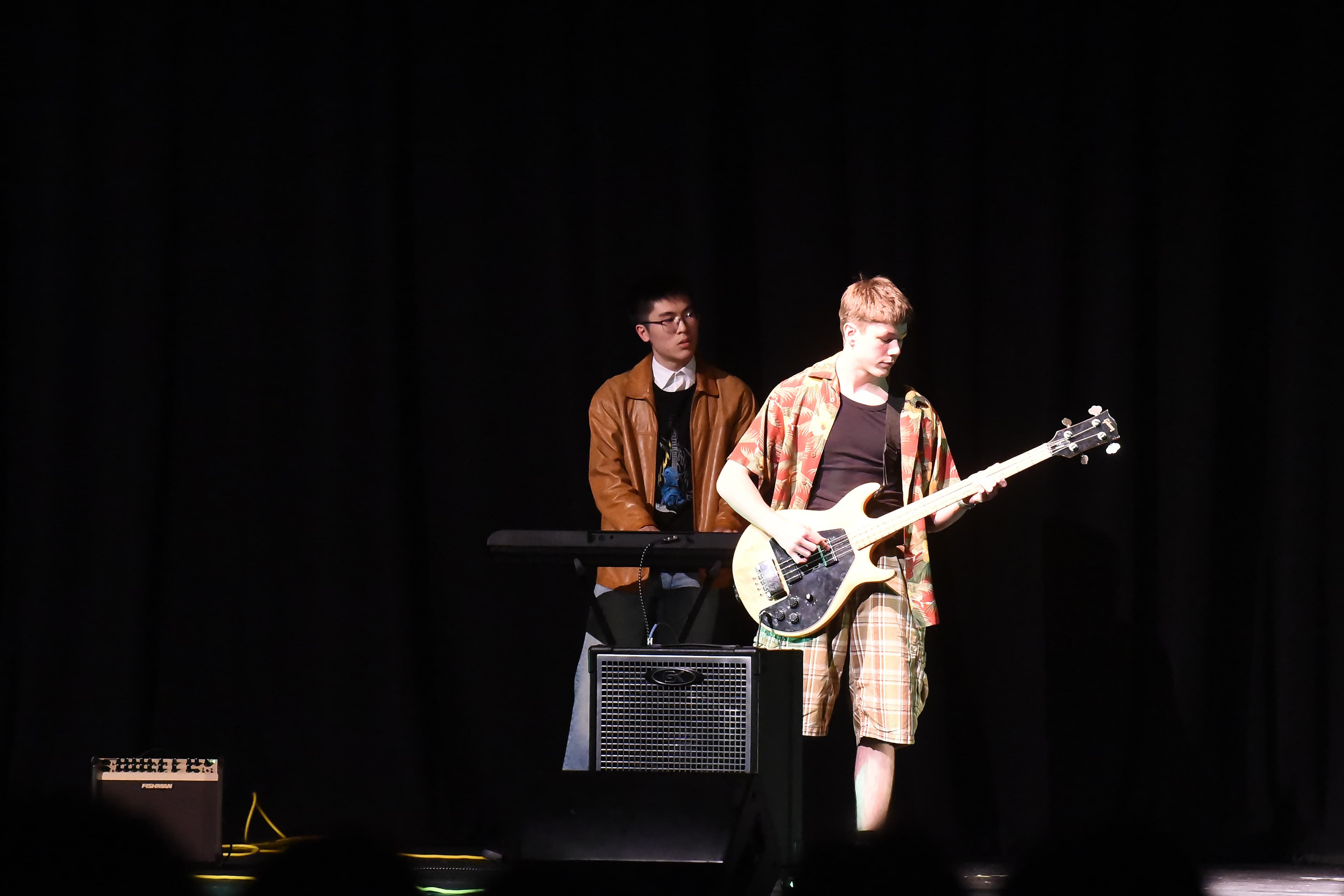 Two student musicians on a dark stage, one playing a keyboard and one playing a bass guitar.