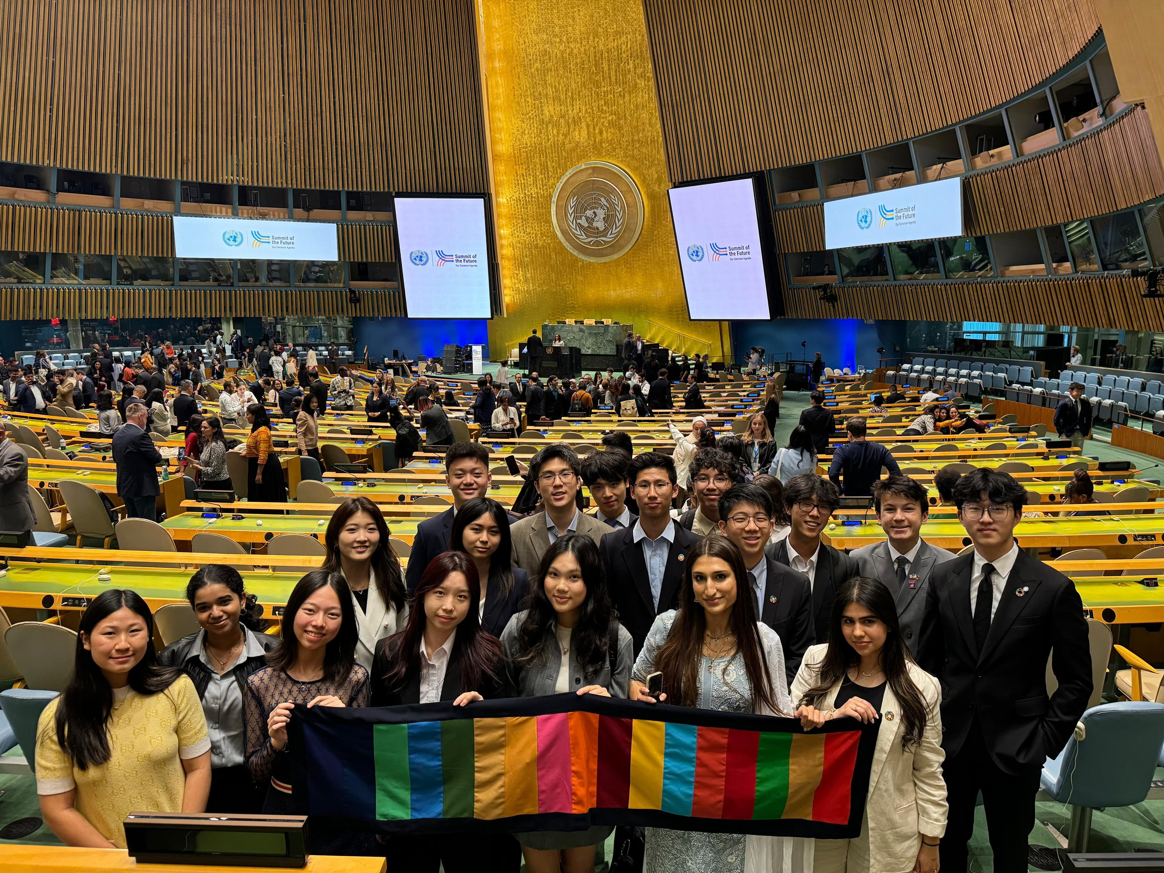 A group of students in formal dress holding a striped flag in the United Nations General Assembly hall, with Summit of the Future screens behind them.